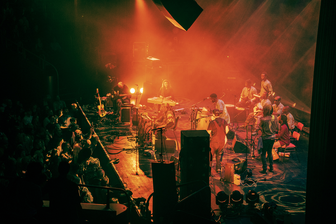 A photo of Lonnie Holley on stage, photographed from the side with musicians behind and on the side, bather in orange lights